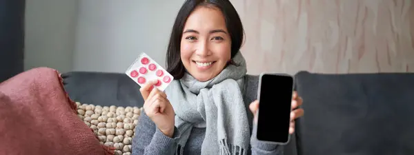 風邪をひいた幸せなアジアの女性は スマートフォンの画面や薬を示し 電話でオンライン医師のアプリ Gpや医療クリニックをお勧めします 薬を保持し インフルエンザで家に滞在 — ストック写真