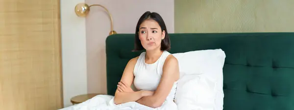 stock image Portrait of asian girl in bed, looking complicated and worried, cross arms on chest, frowning with uneasy, thinking face expression.