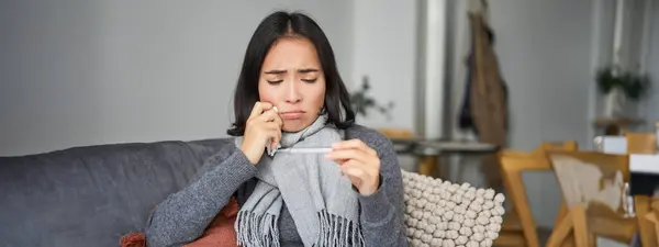 Girl looks upset at thermometer with high temperature, has fever, staying at home ill, feeling unwell, sitting in scarf at home.