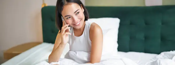 Smiling korean girl talks on mobile phone and lying in bed. Cute woman answers telephone call, holds smartphone, relaxing in her bed.
