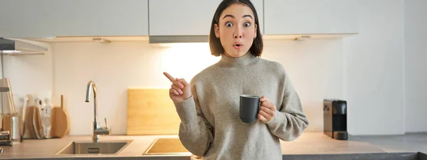 Portret Uśmiechniętej Azjatki Stojącej Kuchni Pijącej Kawę Kubka Wskazującej Baner — Zdjęcie stockowe