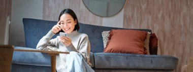 Dikey çekimde, zarif Koreli kadın evinde oturmuş, cep telefonunu kullanıyor, akıllı telefon tutuyor ve gülümsüyor..
