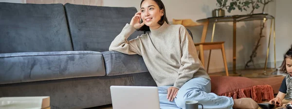Image of smiling asian woman drinking hot tea, holding cup and sitting near laptop on floor, resting at home.