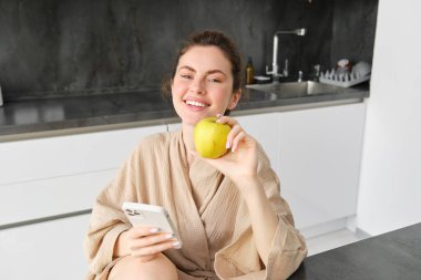 Bornozlu mutlu genç kadının portresini yakın plan çek, mutfakta oturuyor ve cep telefonu kullanıyor, bir elma tutuyor, internetten meyve ve sebze sipariş ediyor, market teslimatı için akıllı telefon uygulaması kullanıyor..