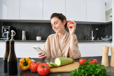 Akıllı telefon uygulamasından alışveriş yapan, elinde domates olan sebze doğrayan genç bir kadının portresi. Tarifi internetten arayan kız, cep telefonu kullanıyor.