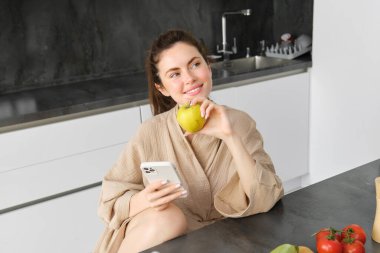 Bornozlu çekici bir genç kadın resmi, mutfakta oturuyor, yeşil elma yiyor ve cep telefonuna bakıyor, akıllı telefon uygulaması kullanıyor, uygulamadan yemek sipariş ediyor..