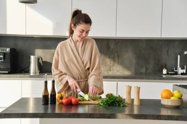 Kahvaltı hazırlayan kadın portresi, salata için sebze doğrama, yemek ve bıçak kullanma, mutfakta dikilme ve bornoz giyme.