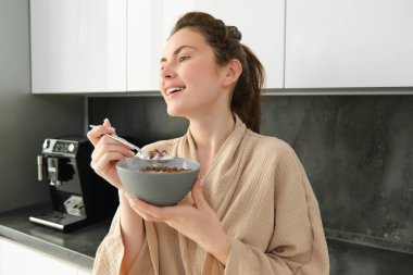 Güzel bir esmer kadın kahvaltısını yapıyor, mutfağın yanında duruyor ve elinde sütle mısır gevreği kasesi tutuyor, sabahının keyfini çıkarıyor, rahat bir bornoz giyiyor..