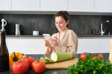 Bornozlu bir kadının portresi mutfakta akıllı telefonuyla oturuyor, yemek pişiriyor, sosyal medyada yemek tarifini izliyor, yemek hazırlamak için video dersi veriyor..
