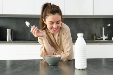 Mutlu genç bir kadının portresi mutfak atölyesine dayanıyor ve mısır gevreği yiyor, önünde süt ve kase yiyor, kahvaltısını yapıyor, bornoz giyiyor..