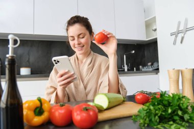 Genç bir kadın mobil uygulamadan alışveriş siparişi veriyor. Bornozlu kız mutfakta sebzelerle oturuyor, yemek tarifi arıyor, akıllı telefon uygulaması kullanıyor..