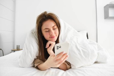Akıllı telefonlu güzel esmer kadının portresi, battaniyenin altında yatağında dinleniyor ve gülümsüyor, cep telefonu uygulamasından sosyal medyayı kontrol ediyor, kendini yorgana sarıp tembelliğin tadını çıkarıyor..