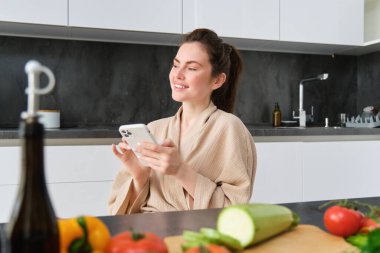 Akıllı telefonlu güzel bir genç kadının portresi, mutfakta oturuyor, yemek pişiriyor, sebzeleri doğruyor. Cep telefonundan yemek tarifini izleyen kız..