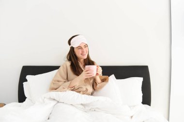 Güzel bir kadının portresi, uyku tulumu ve pijama giymiş, yatakta kahve içen, aydınlık ve rahatlatıcı sabahının tadını çıkaran..