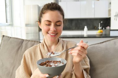 Muhteşem esmer kadın portresi kanepede oturuyor, kahvaltısını evde yapıyor, gevrek yerken gülümsüyor, kase ve kaşık tutuyor ve gülüyor..