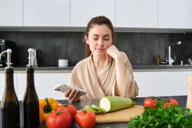 Akıllı telefondan yemek tarifi izleyen bir kadın, mutfakta sebzeler ve kesim tahtasıyla oturuyor, akşam yemeği hazırlıyor, sağlıklı salata yapıyor, evde yemek pişiriyor, bornoz giyiyor..
