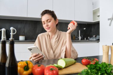 Genç, güzel bir kadının resmi, elinde domates, mutfakta akıllı telefonuyla oturması, tezgahta tahta ve sebzeleri doğraması, yemek pişirmesi, yemek siparişi vermesi, cep telefonu uygulaması kullanması..