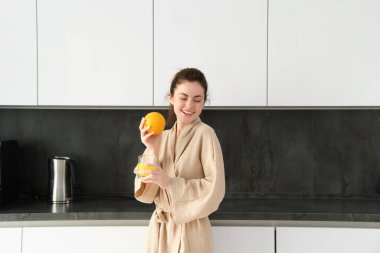 Bornozlu güzel esmer kadının portresi, portakal suyuyla poz veriyor, taze meyve suyu hazırlıyor, sağlıklı bir yaşam sürüyor, sabahına vitamin içeceği ile başlıyor..