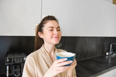 Gülümseyen kadının portresi kahve içer, mutfakta durur ve sabahları lezzetli bir fincan kapuçino içer, kameraya mutlu bakar..