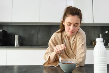 Evde sütle mısır gevreği yiyen, kahvaltı yapan, bornoz giyen, mutfakta tek başına oturan mutlu genç bir kadın..