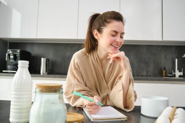 Çekici, genç, neşeli kız mutfakta yemek pişiriyor, para kazanıyor, yemek tarifi kitabını tutuyor, fikirleri var..