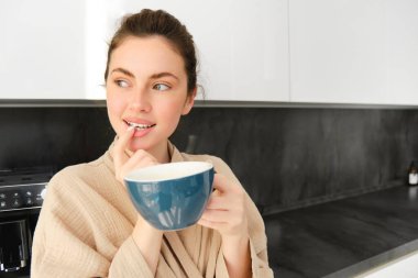 Çekici bir kızın, kahve içen, sabah kapuçinosuyla bardak tutan ve gülümseyen, mutfakta bir kupa lezzetli içki içen, rahat bir bornoz giyen portresi..