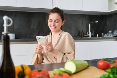 Akıllı telefondan yemek tarifi izleyen bir kadın, mutfakta sebzeler ve kesim tahtasıyla oturuyor, akşam yemeği hazırlıyor, sağlıklı salata yapıyor, evde yemek pişiriyor, bornoz giyiyor..