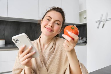 Güzel gülümseyen bir kadının portresi, elinde taze domates, mutfakta akıllı telefonuyla oturmak, internetten sebze siparişi vermek, market alışverişi yapmak, cep telefonu kullanmak..