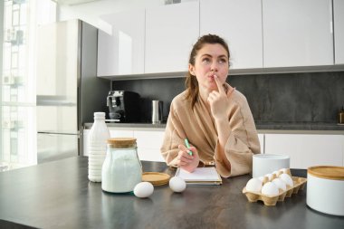 Çekici, genç, neşeli kız mutfakta yemek pişiriyor, para kazanıyor, yemek tarifi kitabını tutuyor, fikirleri var..