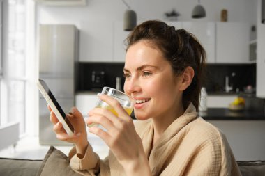 Evde güzel bir kadının portresi, cep telefonu kullanıyor ve bir bardak portakal suyu içiyor..