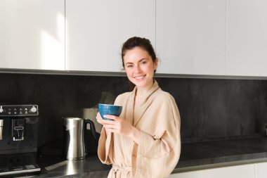 Günlük rutin ve yaşam tarzı. Bornozlu genç bir kadın, mutfakta elinde kahveyle duruyor, çay içiyor, gülümsüyor ve mutlu görünüyor..