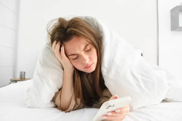 Depressed woman in bed, lying under blanket and looking at phone with sad, complicated face.