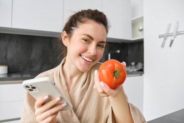 Gülümseyen güzel bir kadın resmi, mutfakta oturuyor, akıllı telefon ve domates tutuyor, mutlu görünüyor, evine yiyecek siparişi veriyor, cep telefonu uygulaması kullanıyor, tarifi için çevrimiçi sebze alıyor..
