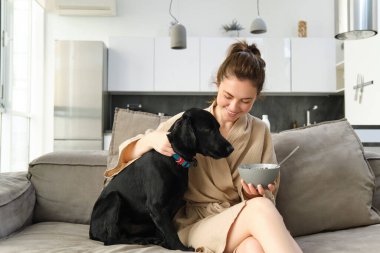 Ev ve yaşam tarzı konsepti. Kanepede oturan, mısır gevreği yiyen, yavru köpeğine sarılan, bornozla kahvaltı yapan güzel bir kadın..