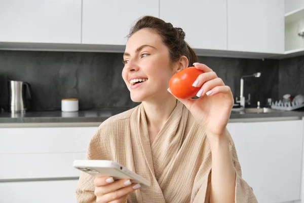 Young woman thinks what to cook, sits in the kitchen with smartphone and tomato in hands, looks aside and smiles, searches recipes on mobile app, orders groceries.