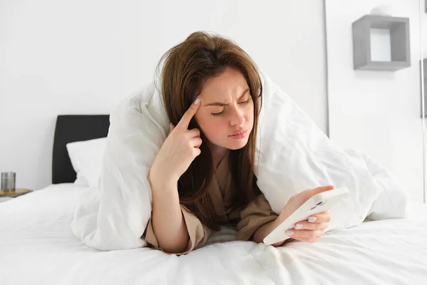 Woman with confused face, waking up in morning and checking her mobile phone, reading message with serious expression, lying in bed under blanket.