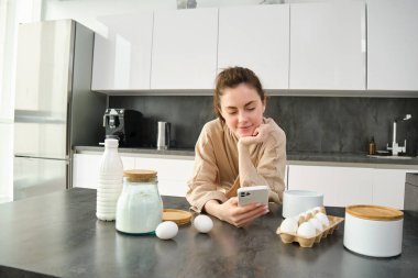 Çekici, genç, neşeli kız mutfakta yemek pişiriyor, para kazanıyor, yemek tarifi kitabını tutuyor, fikirleri var..