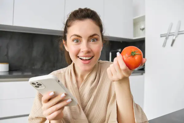 Close up portrait of beautiful young woman in the kitchen, holding tomato and smartphone, buys fresh vegetables online, orders groceries delivery on mobile phone application.