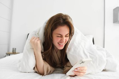 Akıllı telefonlu bir kadın, heyecan duyuyor, cep telefonu okuyor ve tezahürat yapıyor, beyaz battaniyenin altında yatıyor, akıllı telefon kullanıyor ve sabahları mutlu görünüyor..