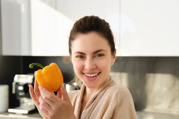 Close up of brunette woman in robe, holding yellow pepper, thinking what to cook, preparing salad or soup, making vegetarian meal with vegetables, standing in kitchen.