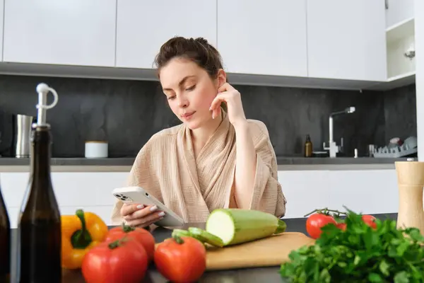 Young woman orders groceries on mobile app. Girl in bathrobe sits in the kitchen with vegetables, looking for recipe to cook dinner, using smartphone application.