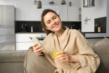 Evde bornoz giyen, cep telefonu tutan, akıllı telefondaki sosyal medya uygulamasını kullanan, portakal suyu içen, kanepede oturan güzel genç bir kadın..