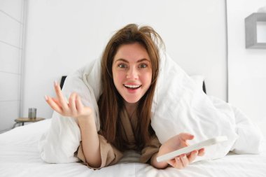 Yatakta yatan ve cep telefonu kullanan etkileyici bir kadın akıllı telefondaki içerikten hayrete düşüyor..