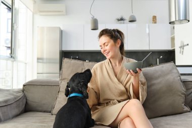 En iyi arkadaşla sıcak sabahlar. Kahvaltıda mısır gevreği yiyen genç bir kadın, kanepede köpeğiyle oturuyor ve gülümsüyor, güne doğru düzgün bir yemekle başlıyor..