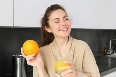 Bornozlu, taze meyve suyu içen, portakal meyvesi gösteren, mutfakta poz veren sağlıklı bir kadın resmi..