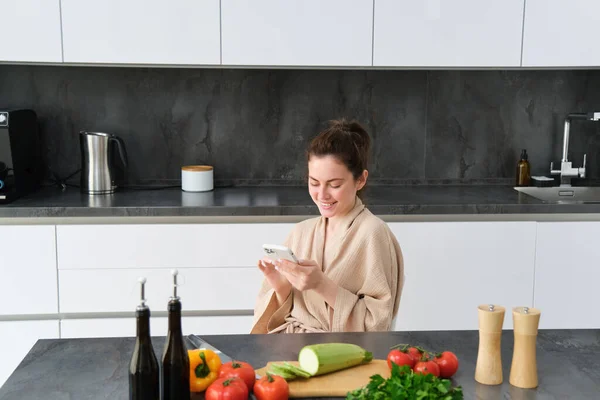 Woman watching recipe on smartphone, sitting in the kitchen with vegetables and chopping board, preparing dinner, healthy salad, cooking at home, wearing bathrobe.