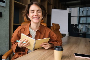 Başlangıç projesinde çalışan, notlar alan, elinde kalem tutan, kağıt okuyan, ofiste oturan neşeli genç bir kadının portresi..