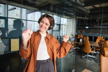 Mutlu genç bayan işverenin portresi, ofiste dans eden iş kadını, kablosuz kulaklıkla müzik dinleyen, akıllı telefonla şarkı söyleyen.