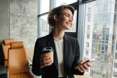 Takım elbiseli, kahve içen, akıllı telefon kullanan, ofiste dikilip pencereden dışarı bakan güzel bir satış elemanı resmi..