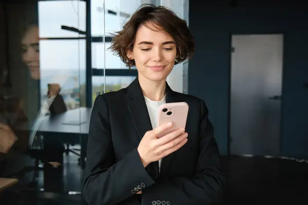 Portrait of corporate woman in suit, standing in office room with smartphone and smiling.
